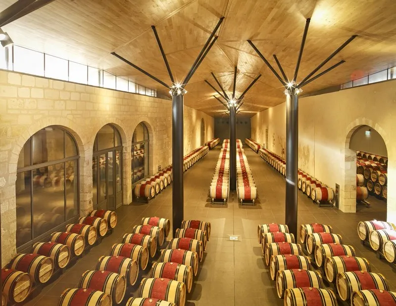 Aging cellar of Château Kirwan in Bordeaux featuring rows of red and white wine barrels lined up beneath a modern architectural ceiling with metal beams.