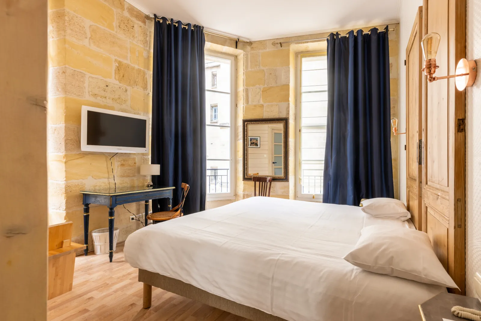 Elegant bedroom at Hôtel de la Tour Intendance in Bordeaux featuring a white bed, golden stone walls, navy blue curtains, black desk, and windows overlooking the street.