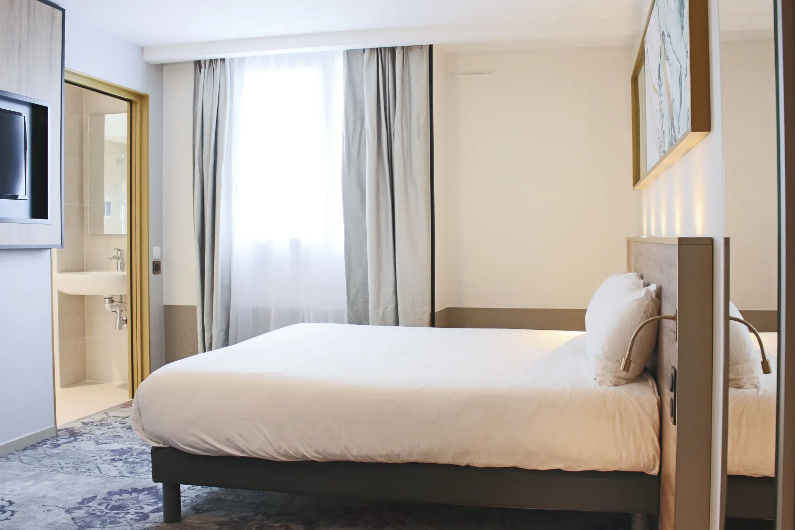 Modern hotel room featuring a queen-size bed, ensuite bathroom, large window with gray curtains, and minimalist decor at Brit Hôtel Bordeaux Aéroport Le Soretel.