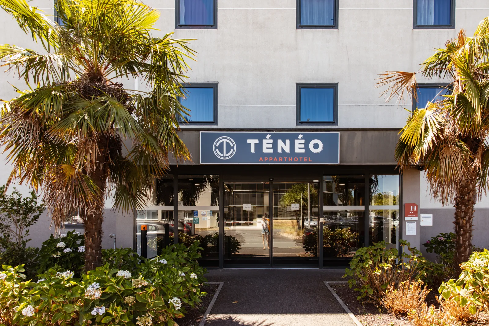 Entrance of Ténéo Aparthotel in Bordeaux-Mérignac featuring modern architecture, tropical palm trees and white flowering plants