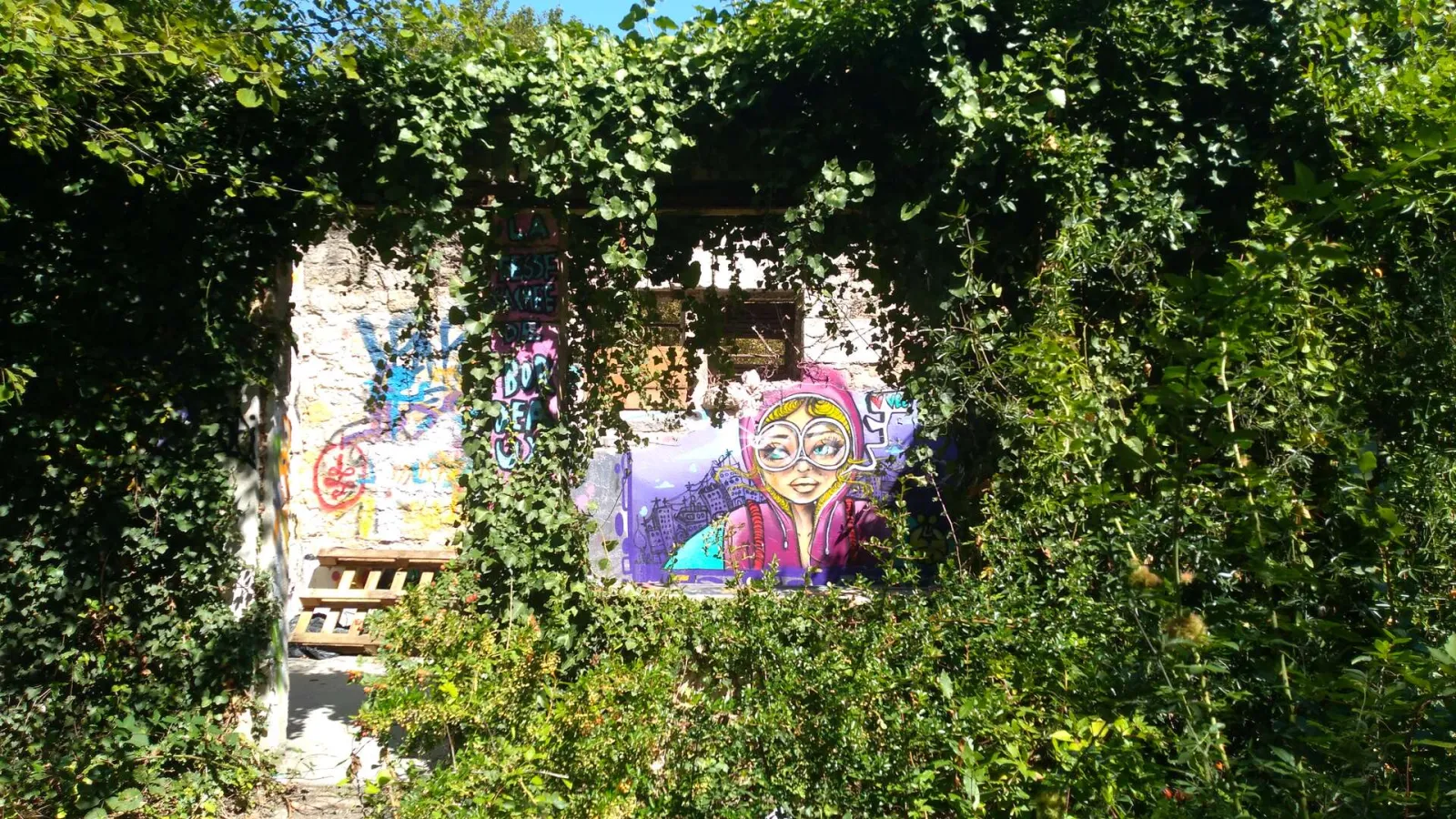 View through a climbing vine archway towards colorful street art murals in the Darwin courtyard, a former military site in Bordeaux.