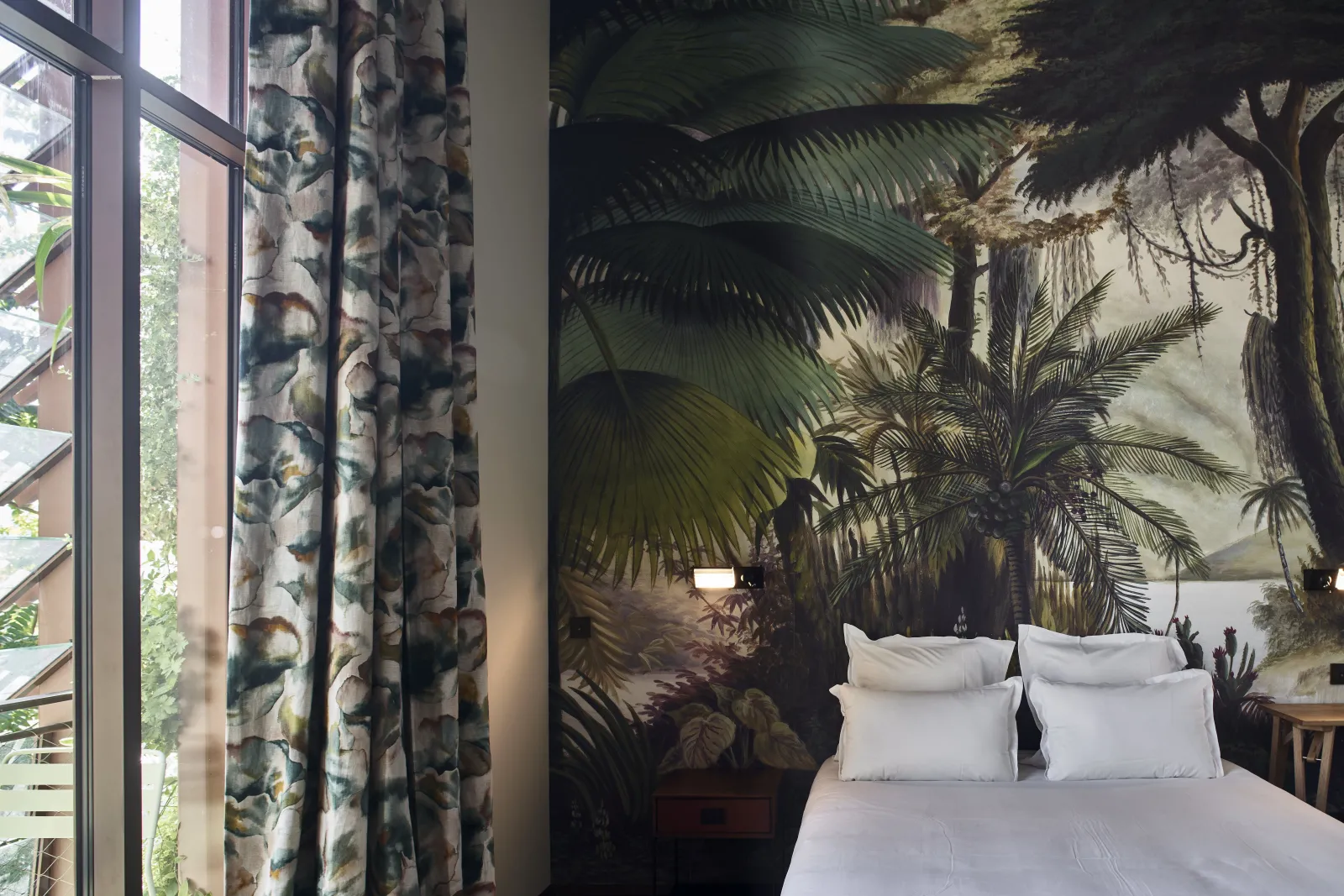 Hotel room at La Zoologie in Bordeaux featuring a white bed, a tropical forest mural with palm trees and lush vegetation, patterned botanical curtains, and a large window overlooking green foliage.