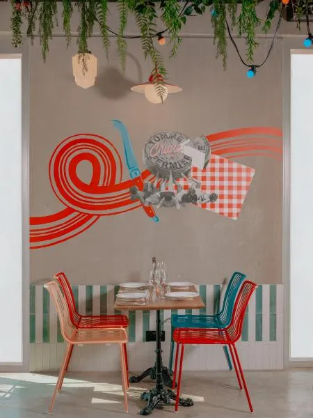 Bordeaux artistic wall installation featuring abstract red and blue patterns, small table with two colorful chairs, hanging plants and French Kiss themed decoration