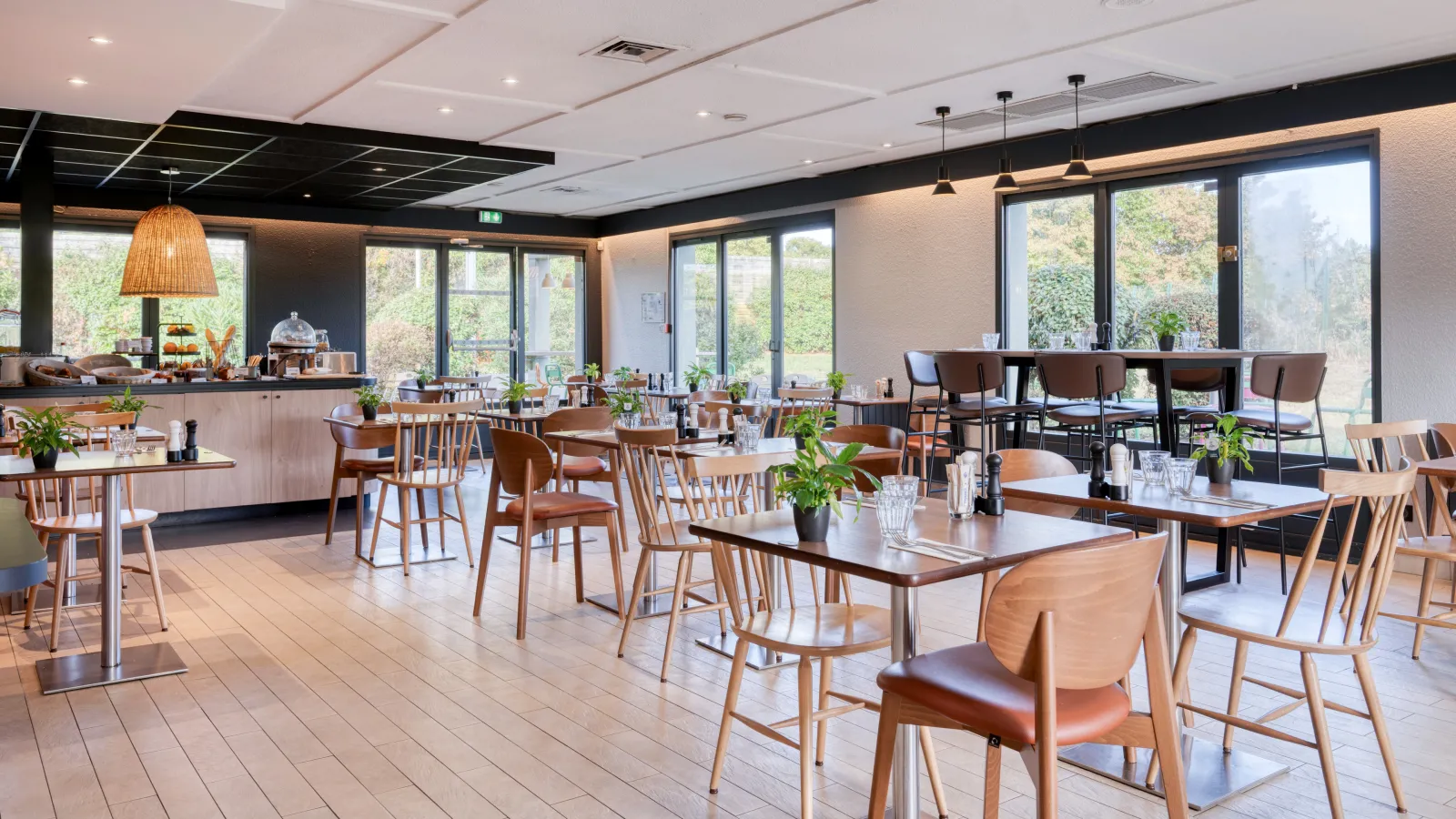 Modern and bright restaurant at Campanile Nature Bordeaux Est featuring wooden tables, designer chairs, large windows overlooking greenery, bar counter, and geometric black and white ceiling.
