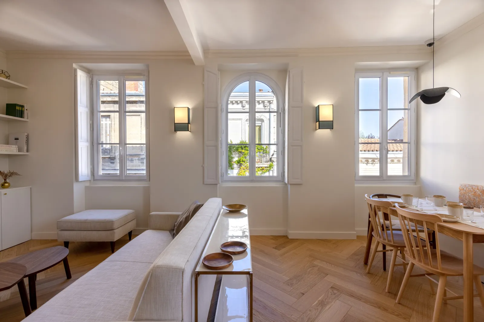 Bright interior of a Haussmann-style apartment in Bordeaux featuring a living room, dining area, three large windows overlooking the city rooftops, modern minimalist furniture and herringbone parquet flooring.