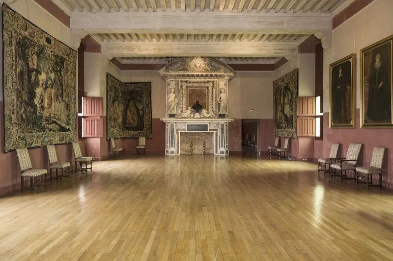 Grand hall of Cadillac Castle featuring light wooden flooring, burgundy walls, ornate white fireplace, ancient tapestries, beige upholstered chairs, and exposed wooden ceiling beams.