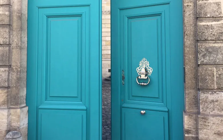 Two turquoise doors with paneled frames and white metal knockers on the stone facade of the small Labottière hotel in Bordeaux.