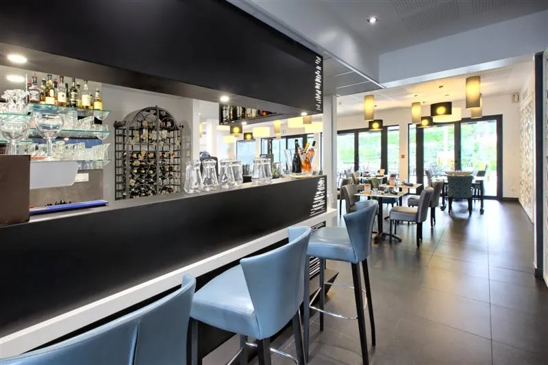 Modern interior of the bar-restaurant at Hôtel Au Comté d'Ornon in Bordeaux, featuring a sleek black counter, blue chairs, suspended yellow lighting, and a bright dining area in the background.