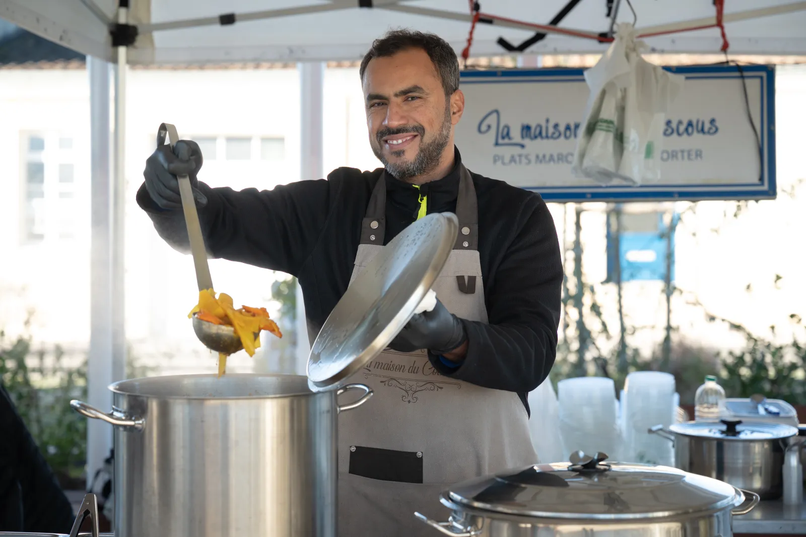 A smiling chef displays a dish prepared in a large pot during a cooking demonstration at the Halle Vettiner in Bouliac Market in Bordeaux, with kitchen utensils and ingredients visible in the background.