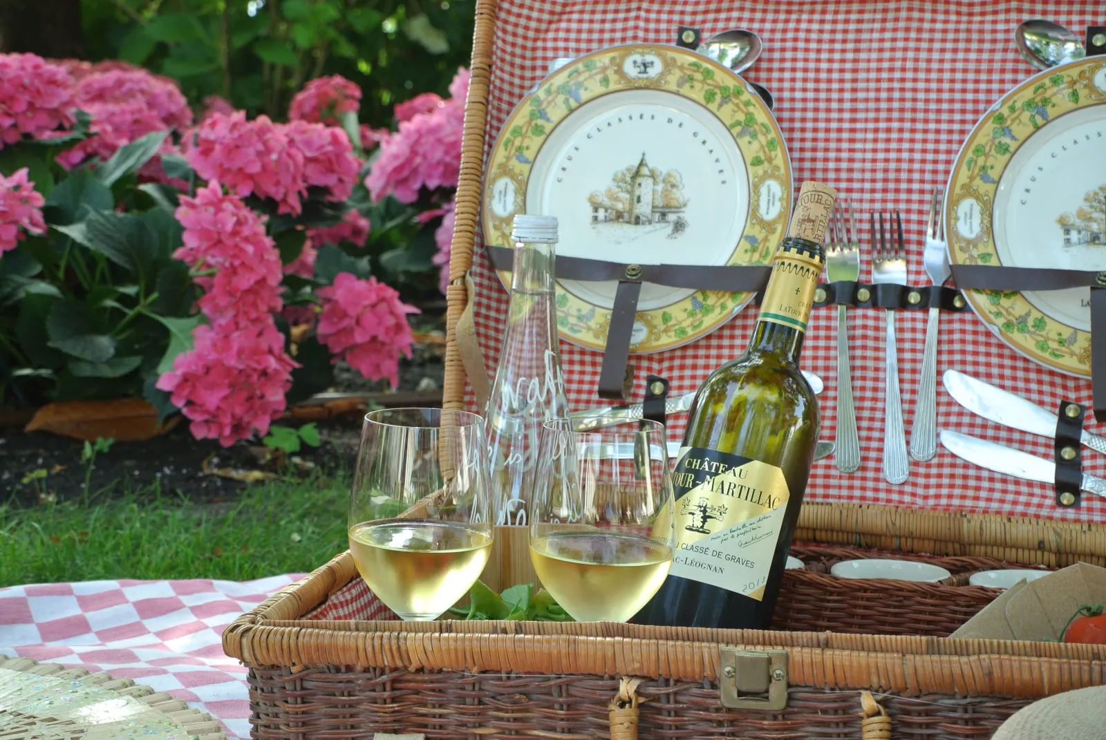 Elegant garden picnic with a wicker basket containing a bottle of Château Latour-Martillac white wine from Bordeaux, two filled glasses, decorative plates with castle motifs, cutlery and red and white checkered napkin, pink flowers in the background.