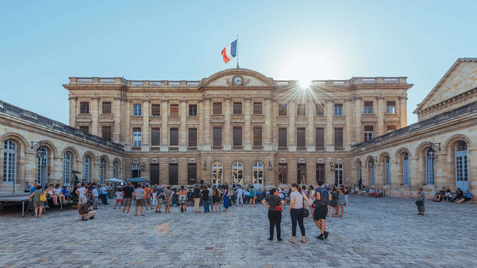 Facade of Palais Rohan in Bordeaux with its paved courtyard, classical stone architecture, French flag on top, and visitors in the courtyard on a sunny day.
