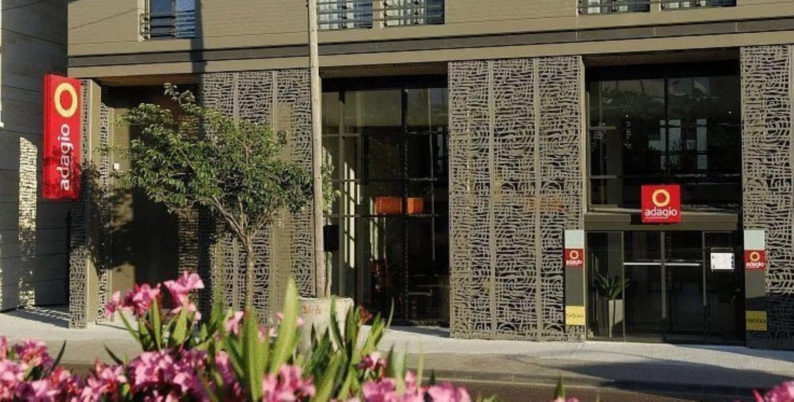 Adagio Bordeaux Gambetta hotel front facade featuring modern design, beige perforated panels, glass entrance doors and pink flowers in foreground