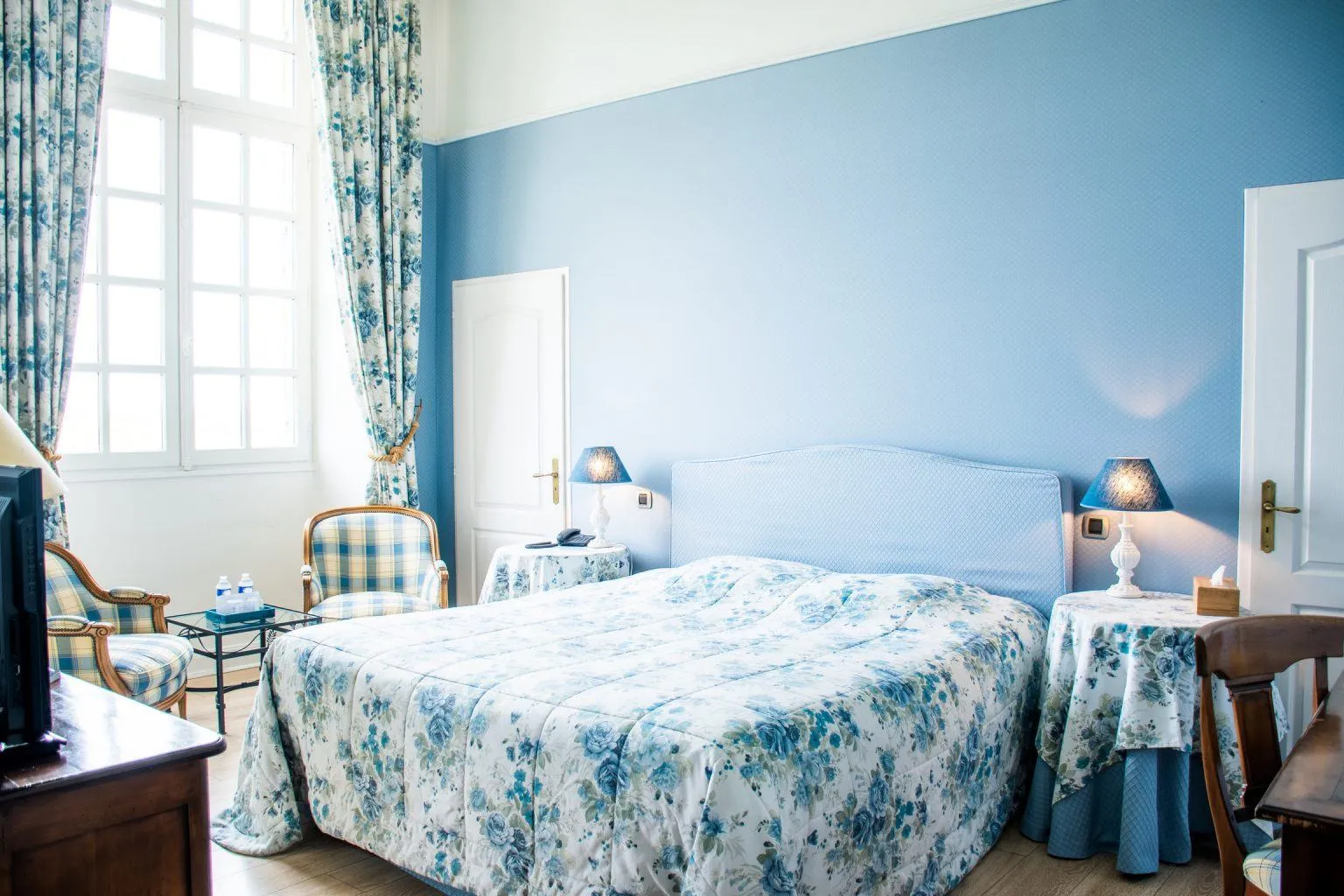 Elegant hotel bedroom with sky blue walls, a double bed with blue and white floral bedspread, matching curtains, wooden furniture, bedside tables with lamps, and a large window.