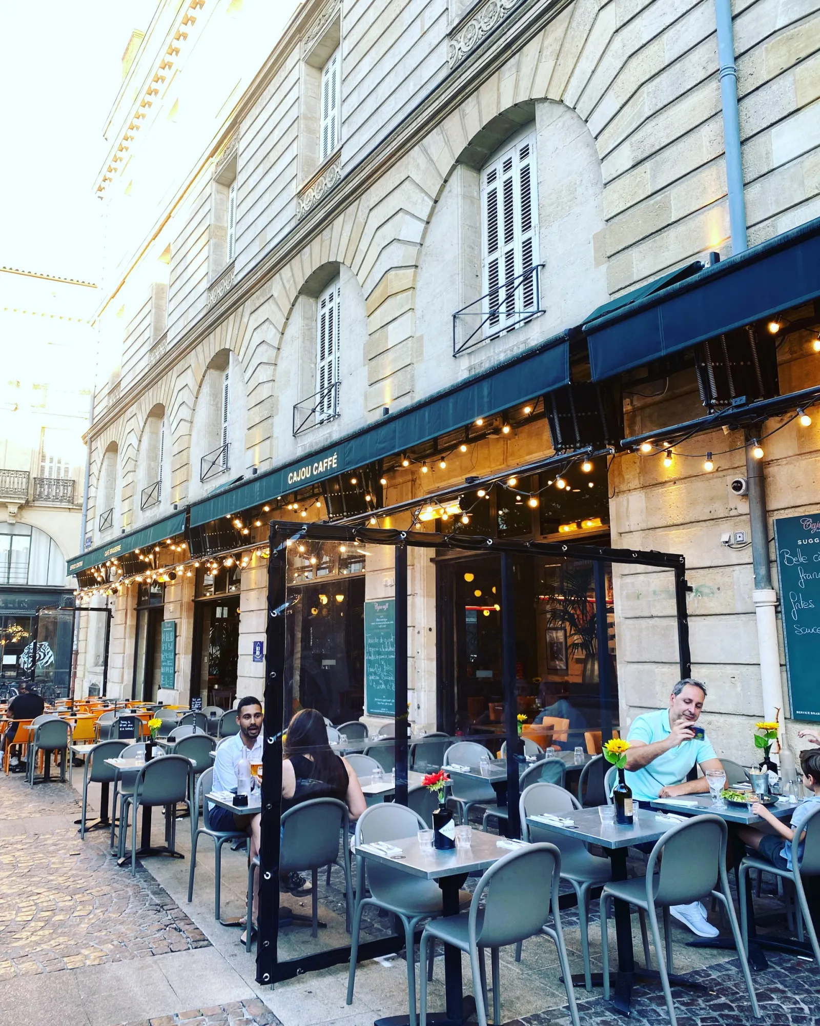 Outdoor seating area of Cajou Caffé café in Bordeaux with customers at tables, Haussmann-style building facade and blue awning.