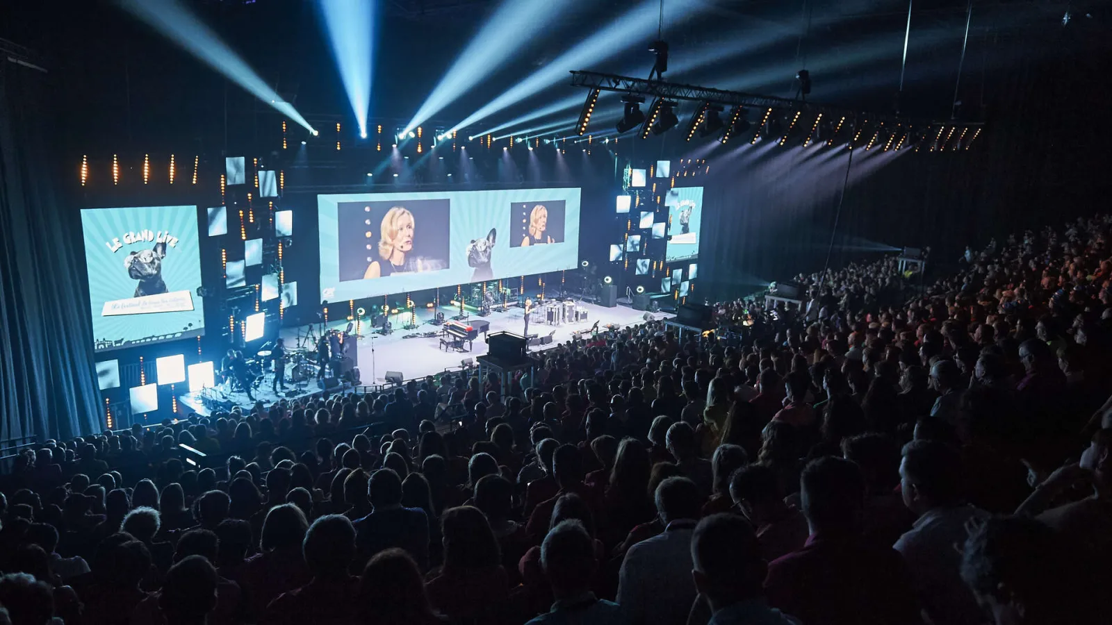 Packed concert hall at Arkea Arena in Floirac with illuminated stage, giant screens and blue spotlights