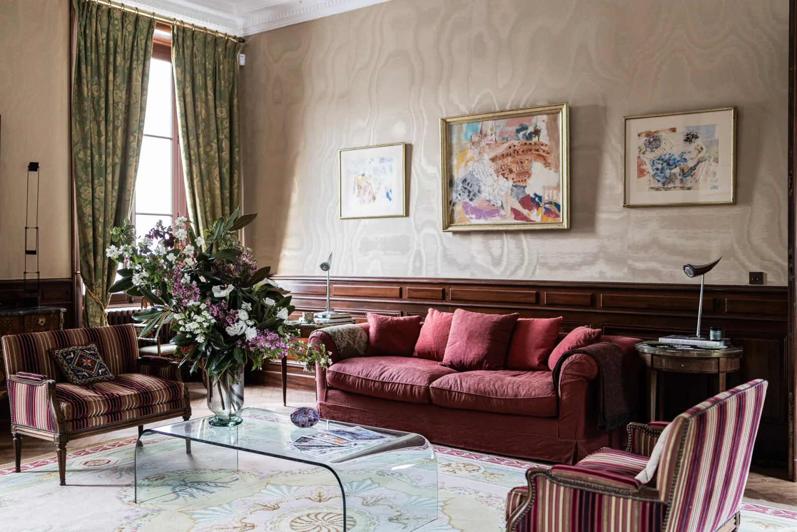 Elegant living room at Château Sénéjac in Bordeaux featuring a burgundy sofa, striped armchairs, a glass coffee table, green curtains, framed artworks, and a flower arrangement by the window.