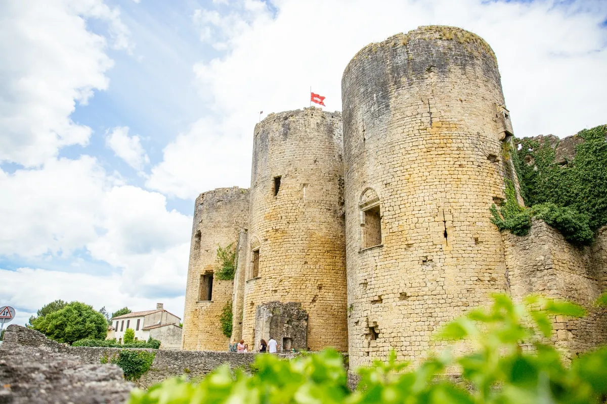 Villandraut Castle near Bordeaux, medieval fortress featuring three cylindrical stone towers, French flag flying, surrounded by lush greenery and fortification walls.