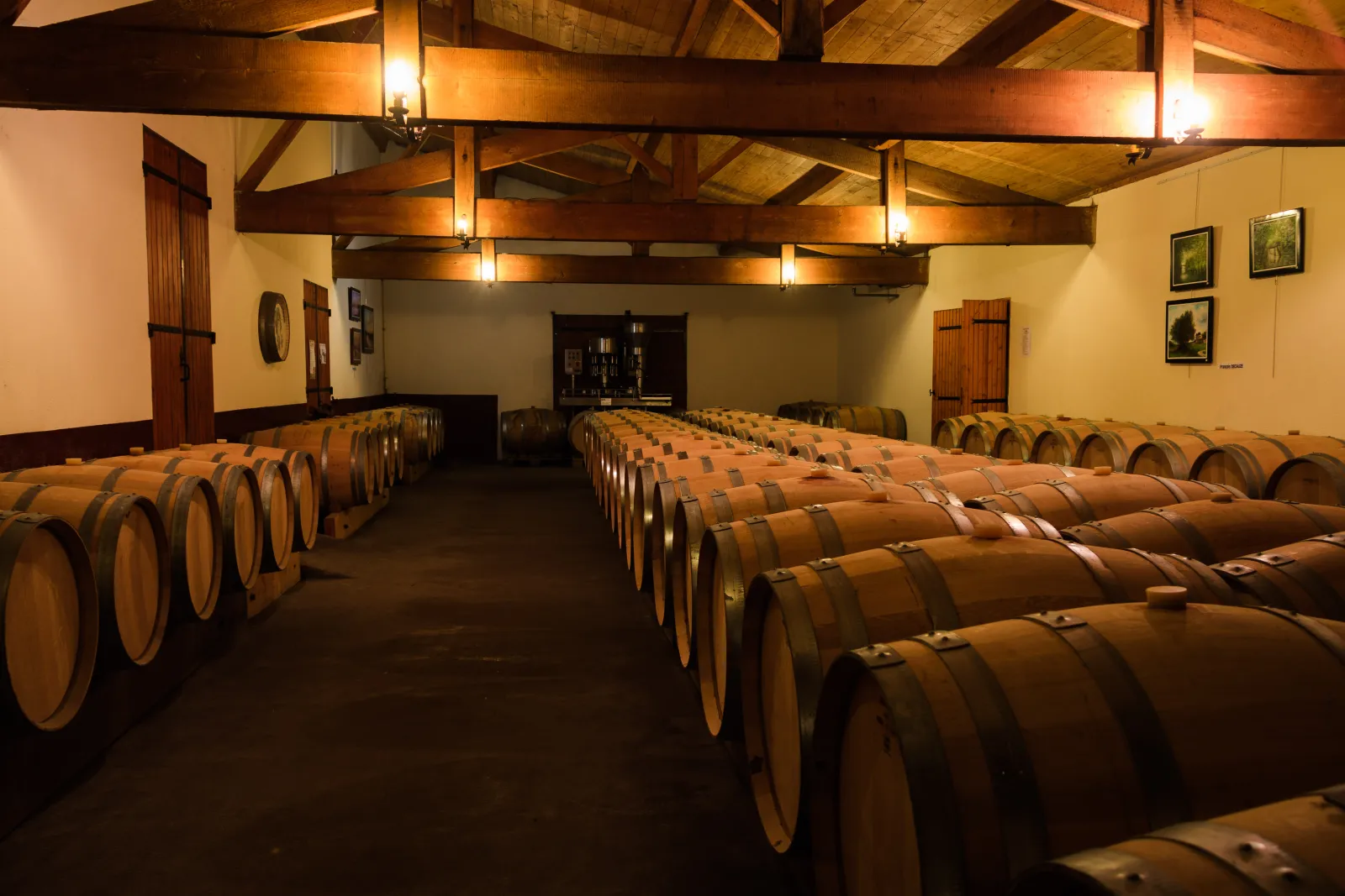 Wine aging cellar at Château Champion in Bordeaux featuring rows of wooden oak barrels arranged symmetrically under exposed timber beams with warm lighting