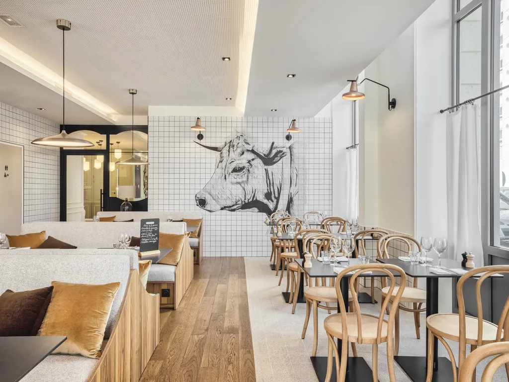 Modern restaurant at Mercure Bordeaux Gare Atlantic hotel featuring wooden tables, bistro chairs, white tiled walls, black and white cow artwork, and large windows overlooking the city.