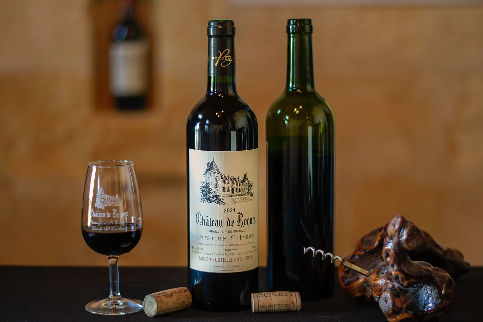 Two bottles of Château de Roques 2021 red wine with a filled wine glass, corkscrew, and tasting accessories on a beige background.