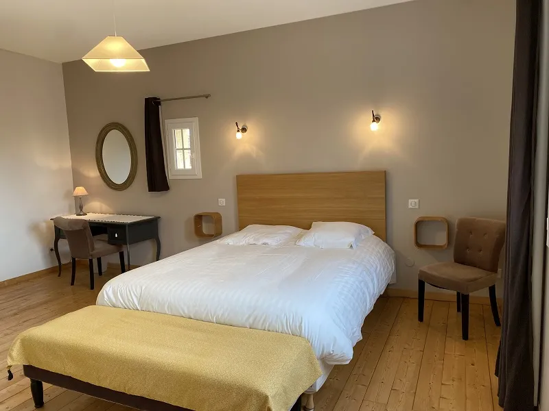 Elegant hotel room featuring a double bed with white linens, wooden headboard, light brown furniture, desk with round mirror, modern chairs, wall sconces and pendant light, light wood flooring at Château La Gontrie in Bordeaux.