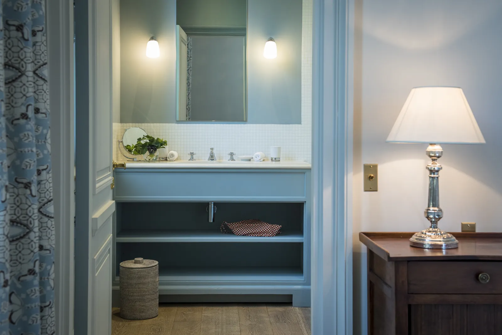 Elegant bathroom featuring a blue vanity, framed mirror with sconces, open shelving with decorative items, woven basket, and a classic floor lamp in a period Bordeaux interior