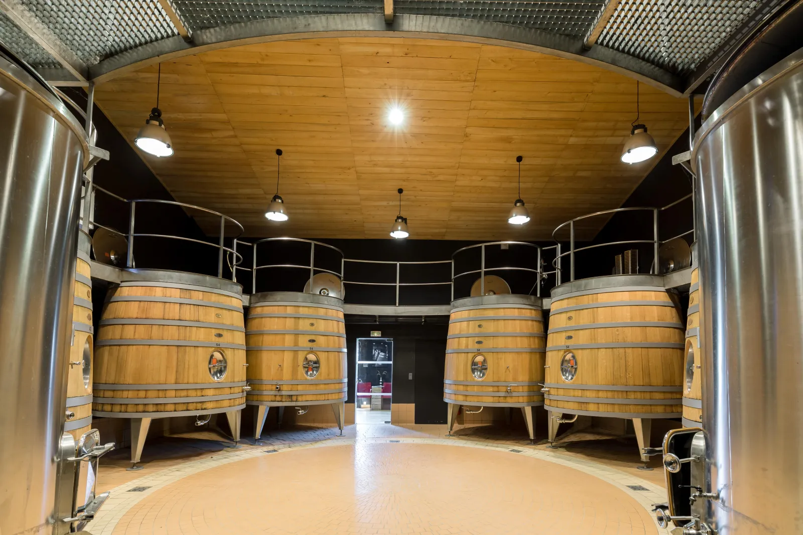Interior of a winery cellar featuring four wooden barrels arranged in a semicircle beneath a wooden ceiling with metal beams and LED lighting, at Château de Candale in Bordeaux.