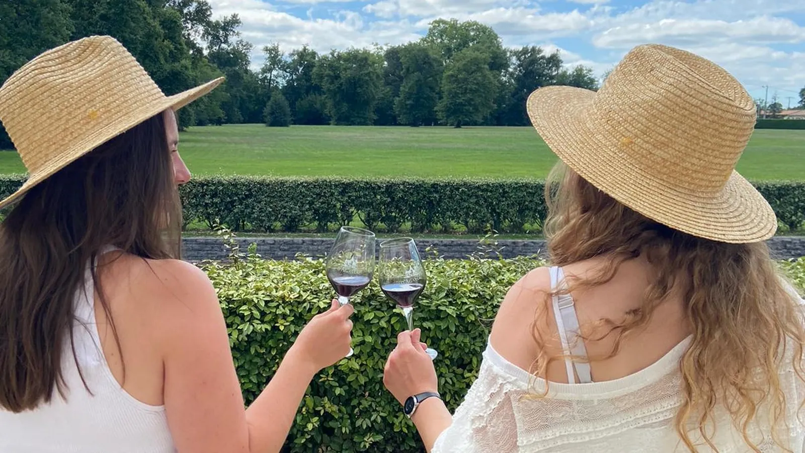Two women wearing straw hats toasting with glasses of red wine in front of vineyards at Château d'Agassac near Bordeaux, with vineyard landscape in the background.