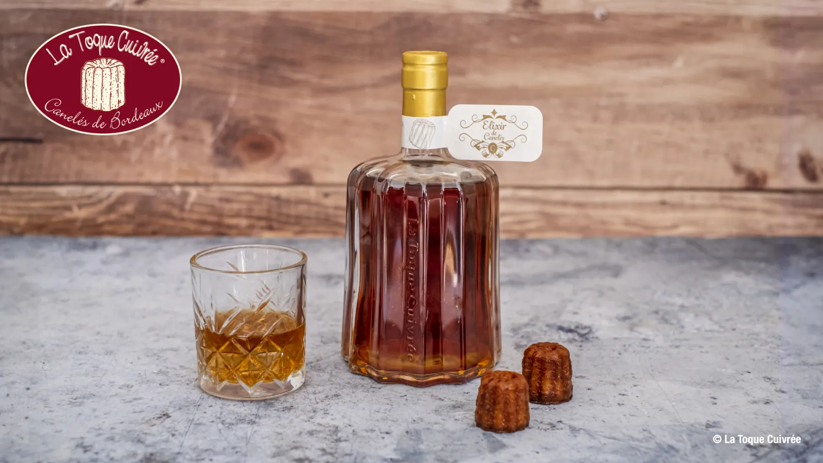 Bottle of elixir from La Toque Cuivrée with a glass of amber liqueur and two Bordeaux canelé pastries on a stone countertop