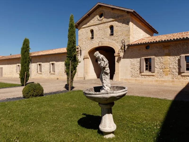 Entrance facade of Château du Lort in Bordeaux featuring a stone portal, cypress trees, ornamental basin and adjoining buildings under clear sky