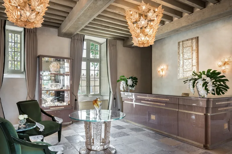 Elegant reception hall of Hôtel Lalique featuring exposed wooden beams, golden crystal chandeliers, solid wood reception desk, green velvet armchair, marble side table, glass doors overlooking the garden, and potted plants.
