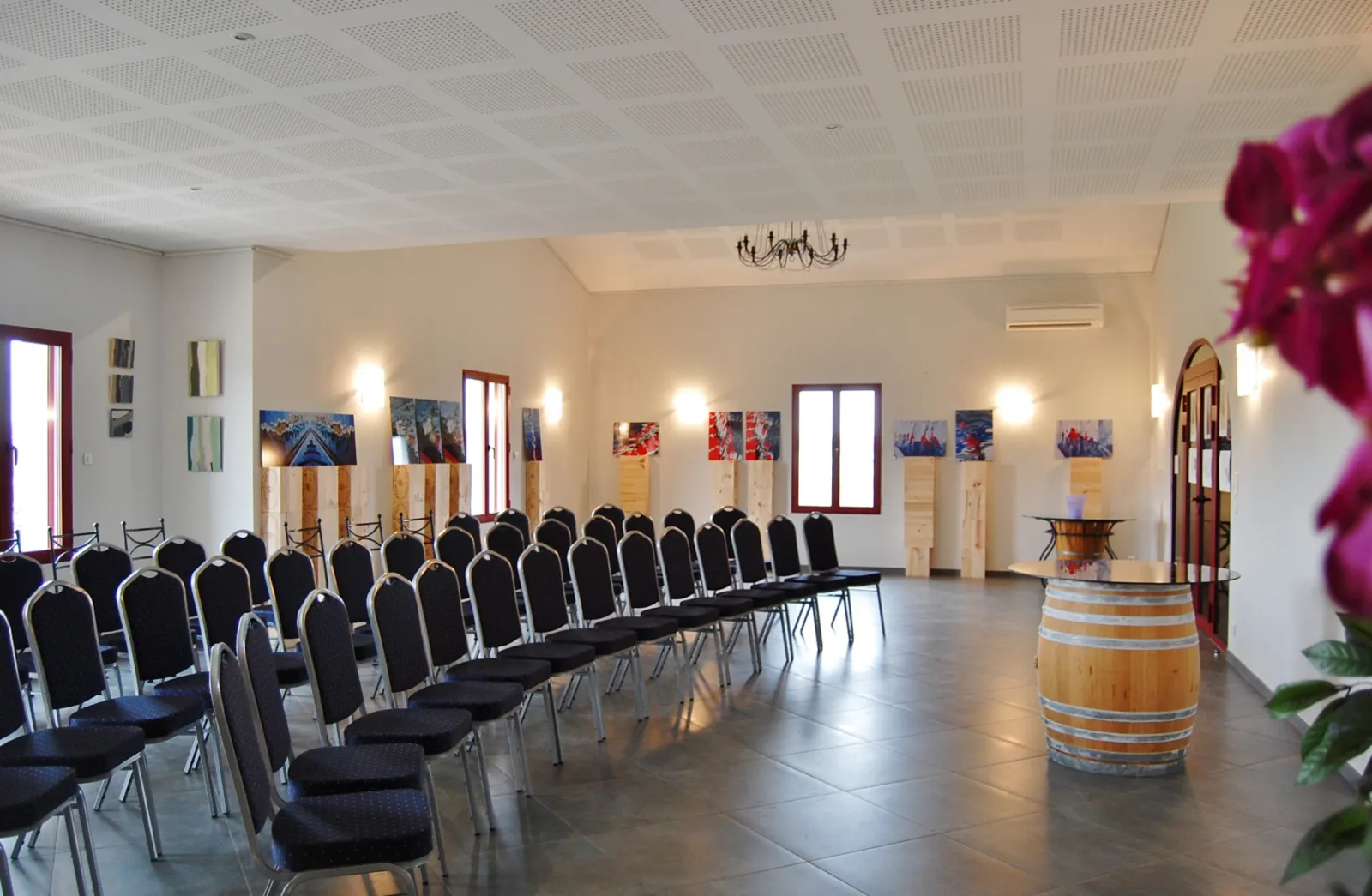 Elegant reception hall at Château Pichon Bellevue in Bordeaux, featuring rows of black chairs, artwork on walls, a decorative wooden barrel, and red flowers in the foreground.