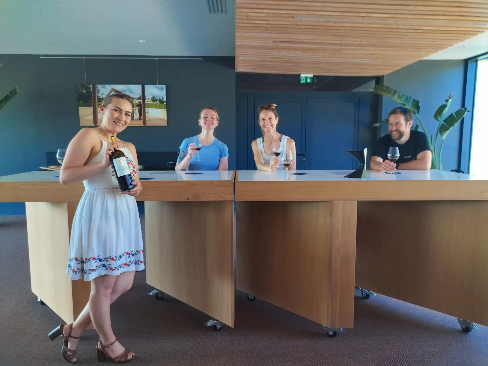 A woman showcases a wine bottle at a counter while four other people watch her in a modern space with blue and wooden decor.
