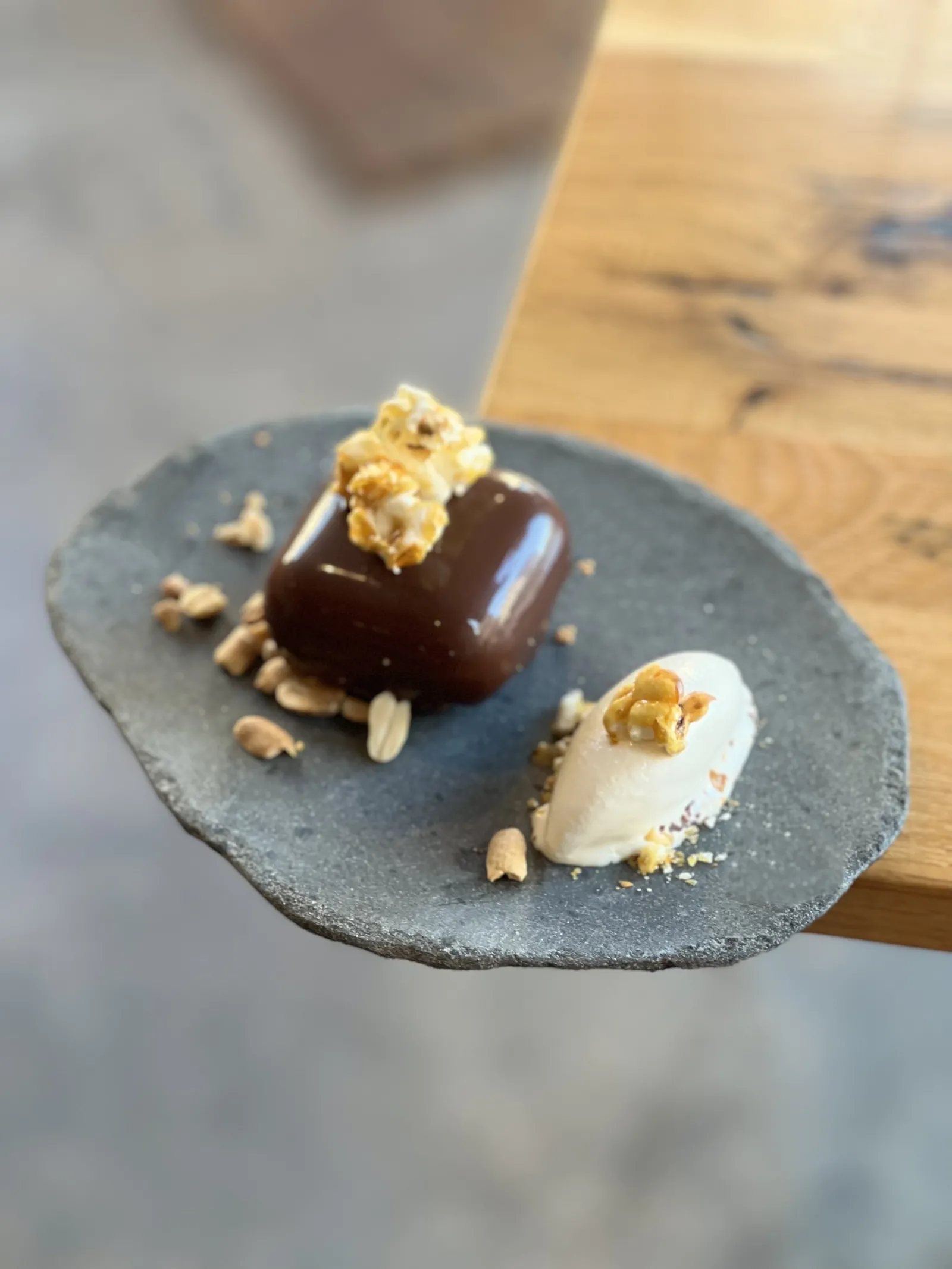 Tasting plate featuring two elegant desserts: a dark chocolate bonbon topped with caramelized popcorn and a white cream quenelle with golden crumbs, presented on a grey slate in a fine dining restaurant in Bordeaux.