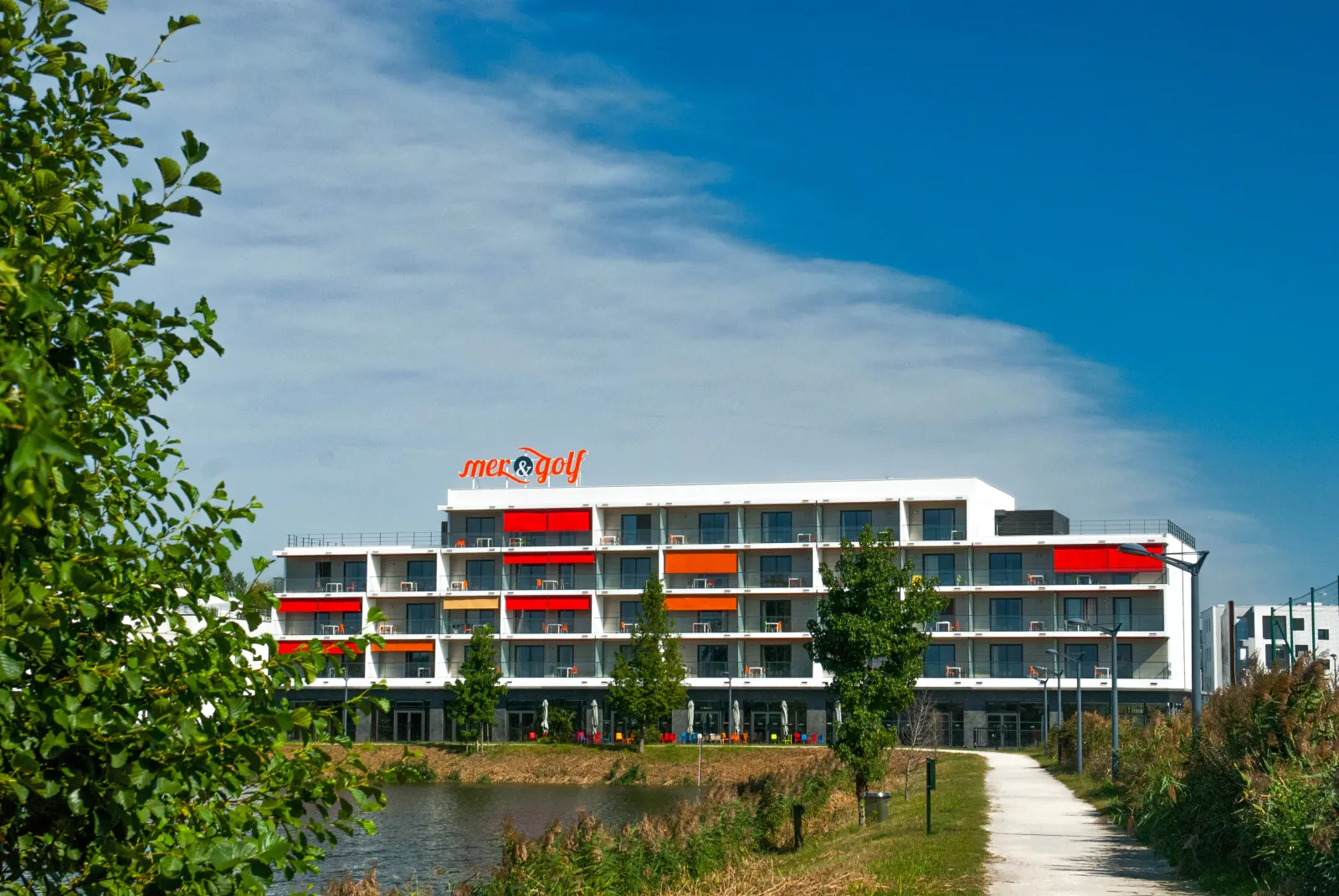 Mer & Golf City Bordeaux Bruges apartment hotel, modern white and red building with balconies overlooking a canal, surrounded by greenery under blue sky.