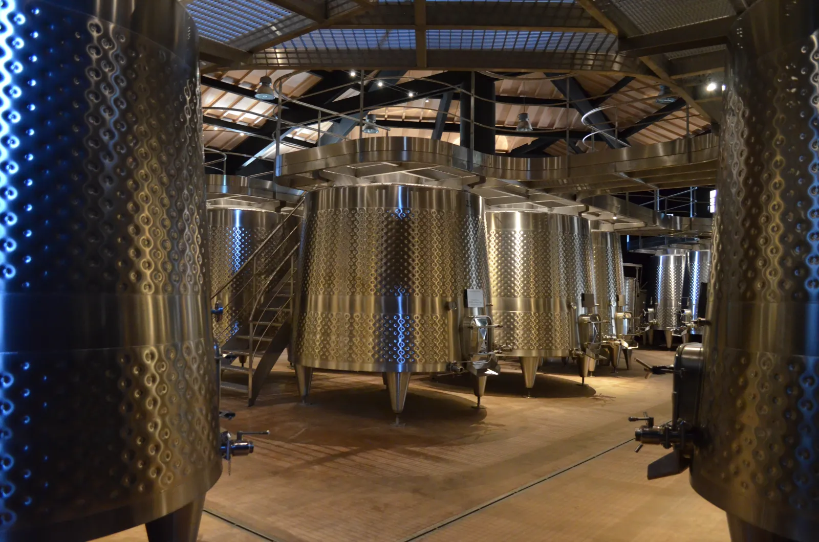 Interior of a modern wine cellar featuring stainless steel fermentation tanks arranged in an industrial building with exposed metal structures and blue-white LED lighting.