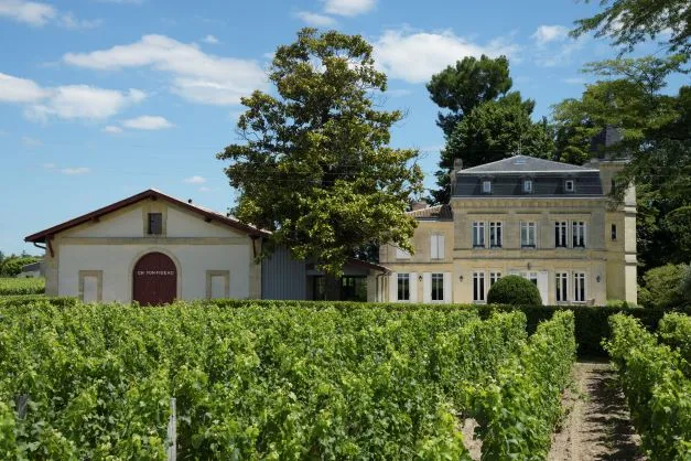 Château Yon-Figeac in Bordeaux featuring a yellow stone mansion, wine estate buildings, and lush vineyards in the foreground against a clear blue sky.