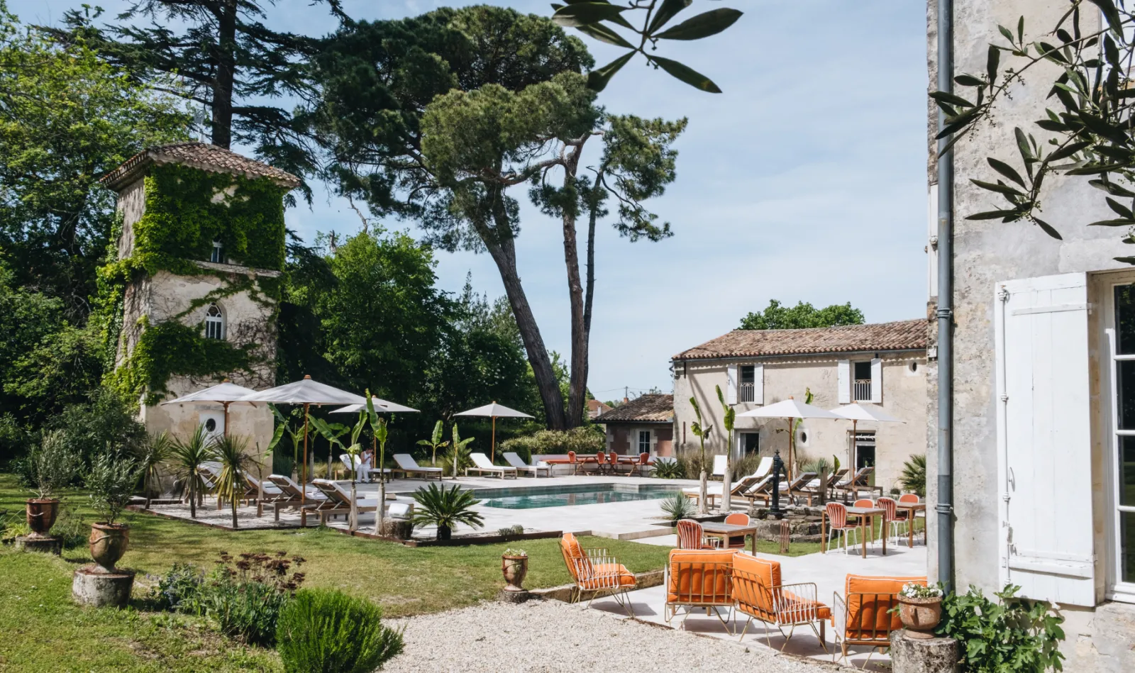 Panoramic view of Château Réal in Bordeaux featuring a swimming pool, orange lounge chairs, stone buildings, manicured gardens and white umbrellas under blue skies