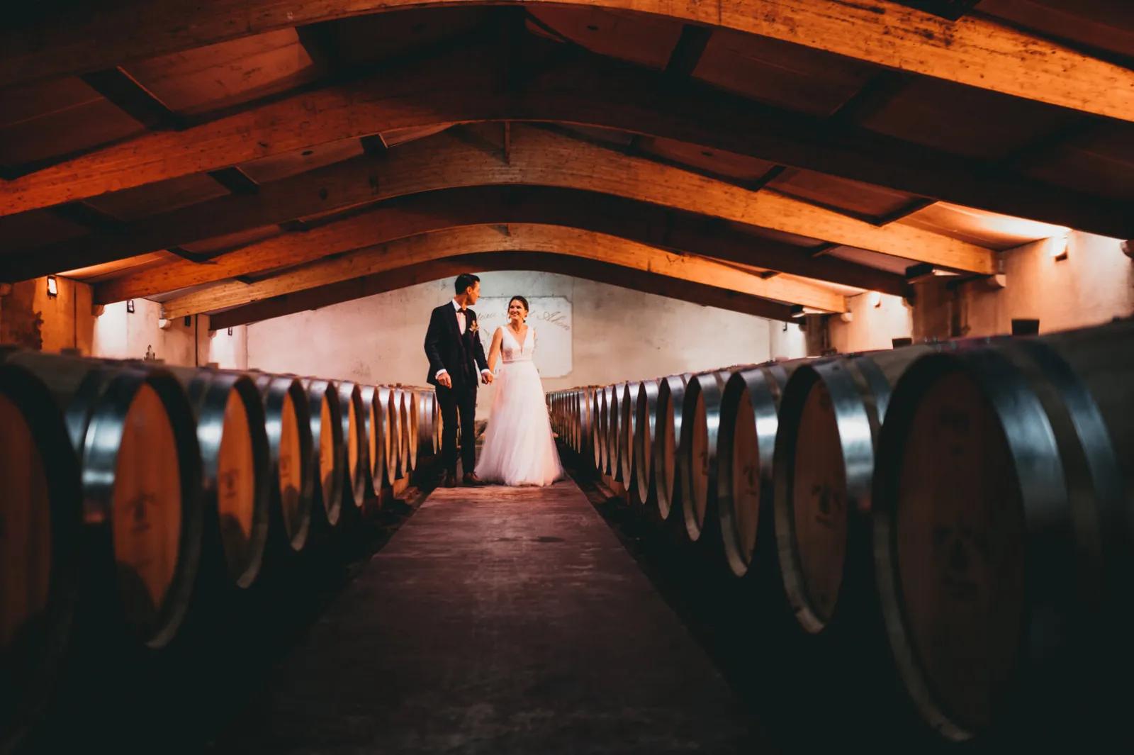 A newlywed couple stands in a barrel cellar at Château Saint Ahon in Bordeaux, surrounded by wine barrels lining both sides.
