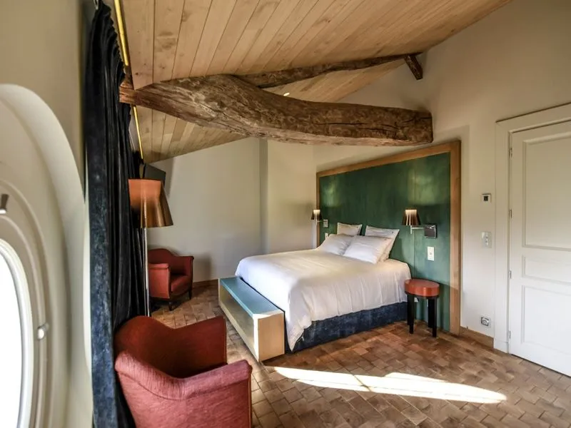 Elegant bedroom at Château Beauregard in Bordeaux featuring a four-poster bed, green headboard, exposed wooden beams, red armchairs, and terracotta tiling.