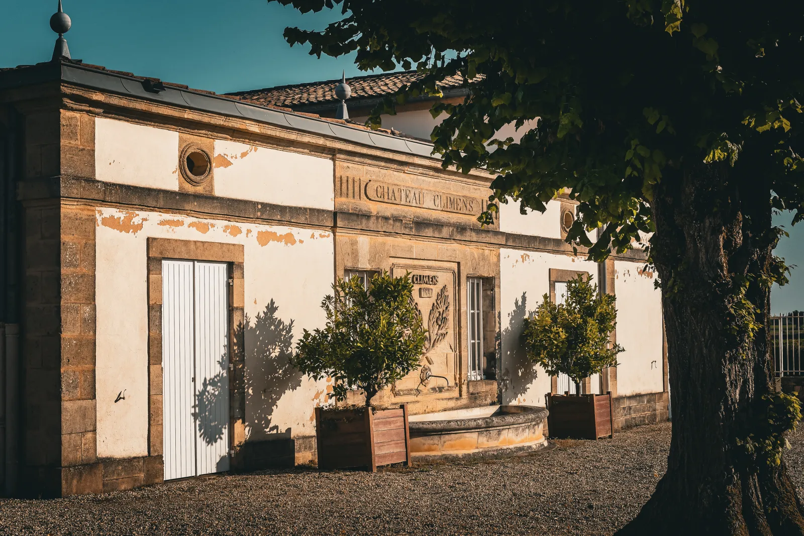 Historic facade of Château Climens in Bordeaux, a beige stone building with white doors, potted plants at the entrance, under clear blue sky.