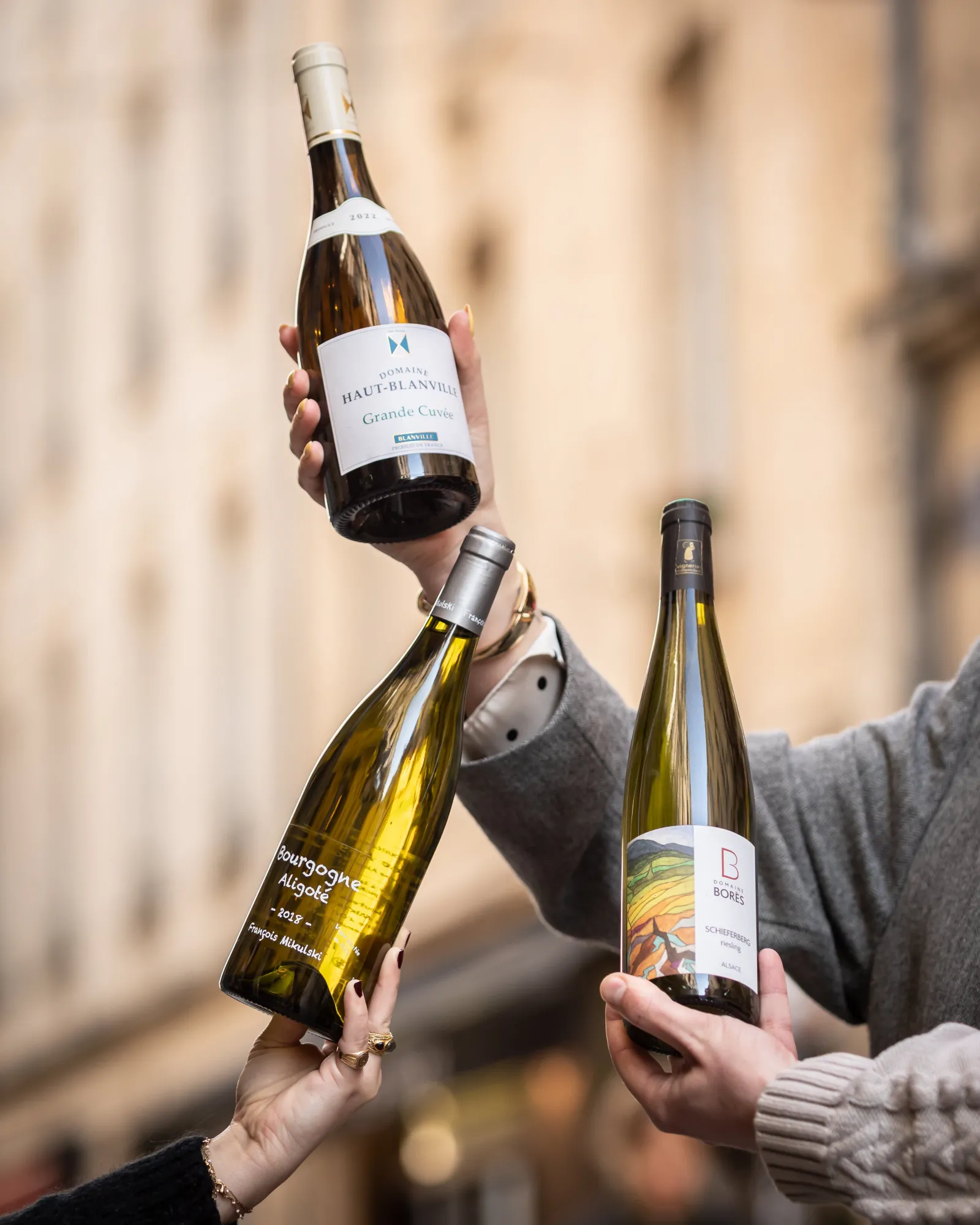 Three people raising glasses of white Bordeaux wine in front of an old building facade, celebrating together at Wine Corner.