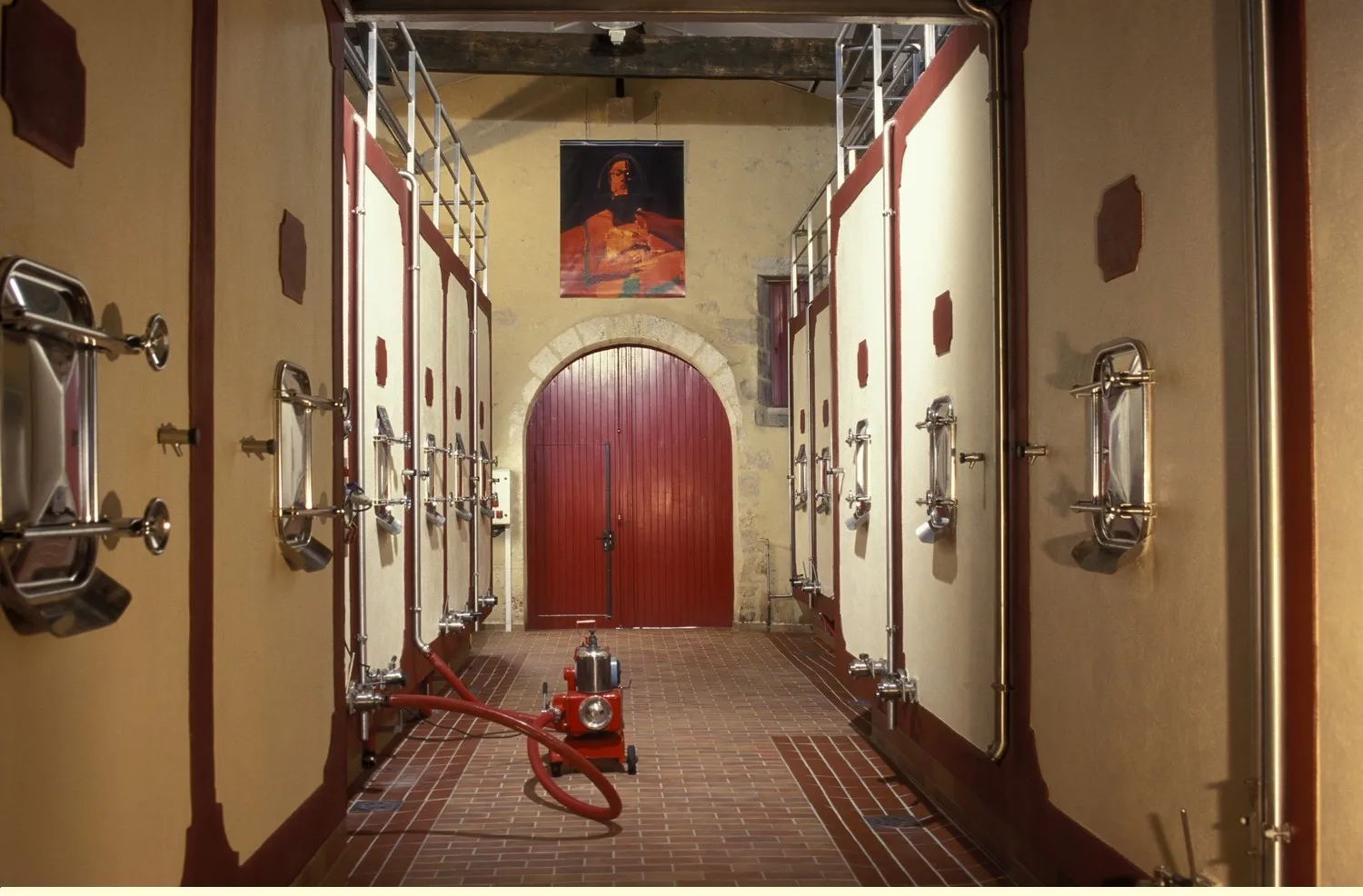 Underground corridor in Château de Pressac, Bordeaux, featuring rows of stainless steel vats, red wooden door at the end and portrait on the wall