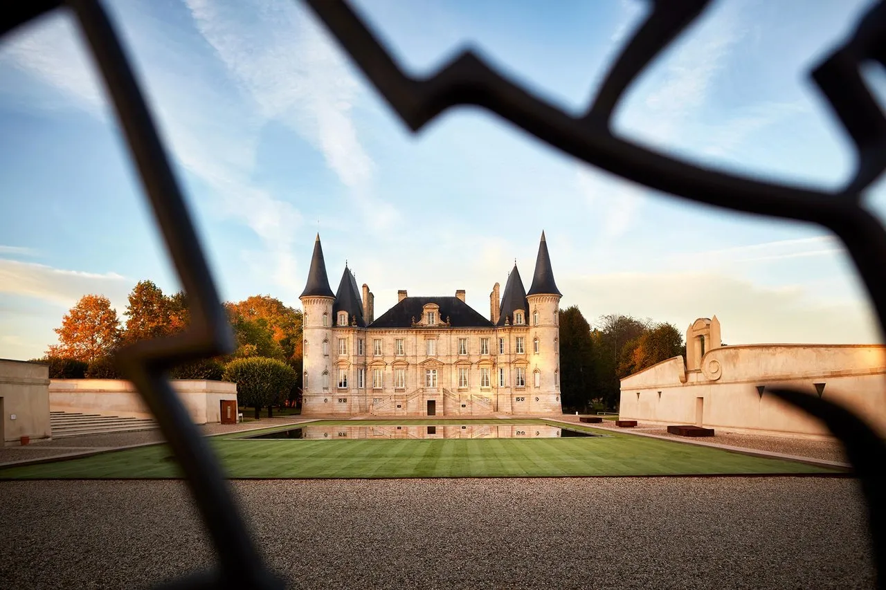 View of Château Pichon Baron in Bordeaux through an ornate wrought iron gate, featuring the main facade with conical towers, manicured lawns, and autumn foliage.