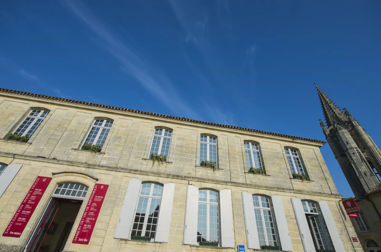 Facade of the Maison du Vin de Saint-Emilion in Bordeaux, a stone building with multi-pane windows, red information banners and a Gothic bell tower visible on the right