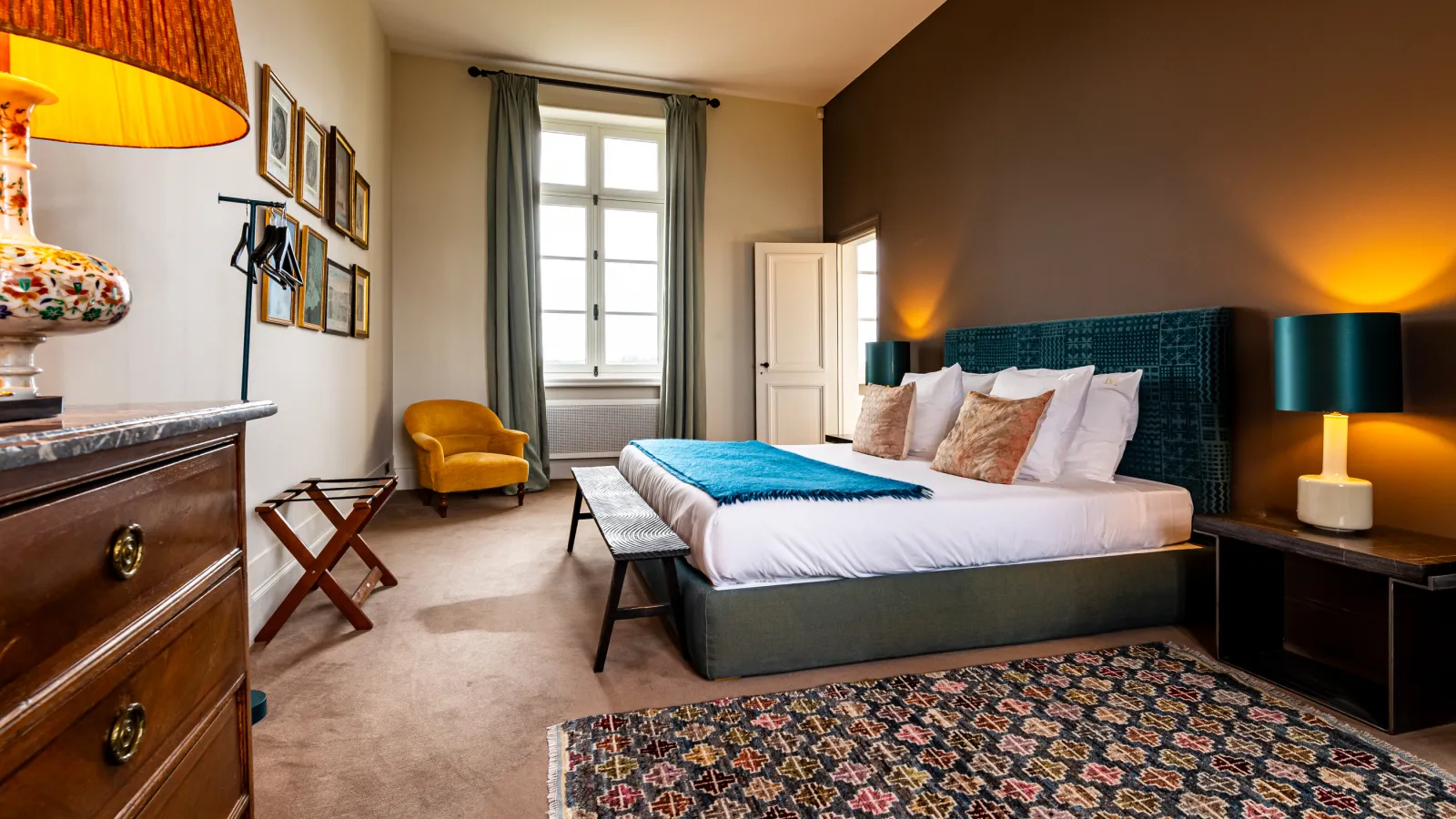 Elegant bedroom at Château Malescasse in Bordeaux featuring a king-size bed with white linens, teal upholstered headboard, wooden furniture, gold lamps, mustard-yellow armchair, window with green curtains, and refined classical decor.