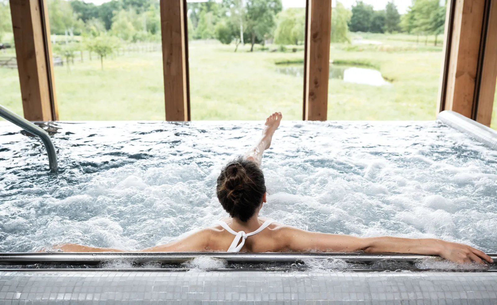 A woman relaxes in a hot tub at Les Sources de Caudalie in Bordeaux, with a view of the surrounding gardens and countryside through large windows.