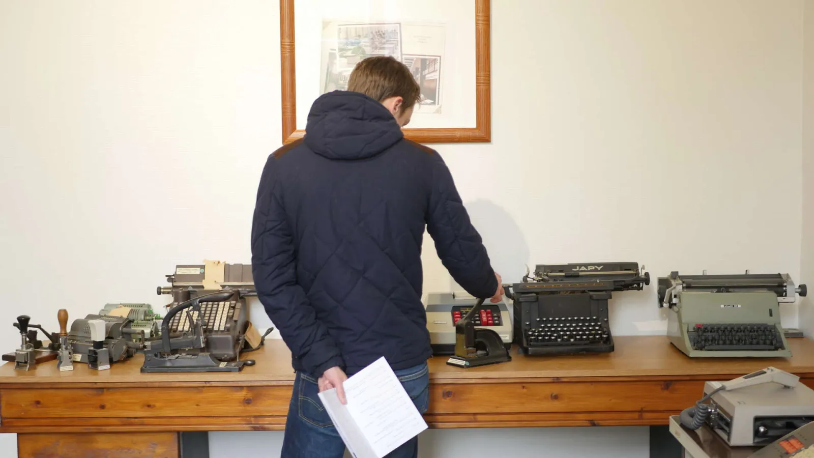 A visitor examines a collection of vintage typewriters displayed on a wooden table in a museum. He holds a document and observes various historical models arranged in a row.