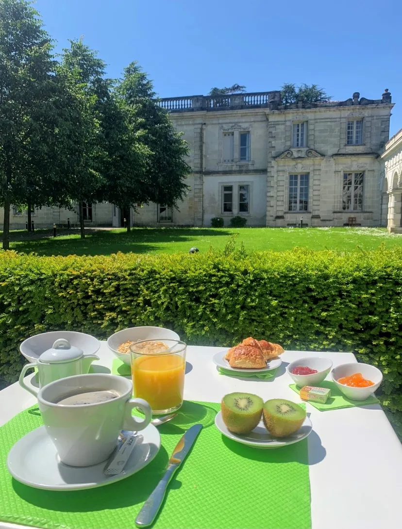Elegant breakfast served on the terrace overlooking Château La Chèze in Bordeaux, with a view of the classic château facade, trees, and manicured gardens.