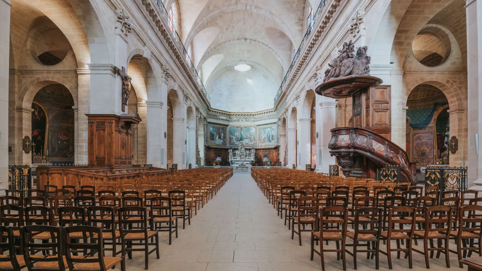 Interior of Notre-Dame Church in Bordeaux featuring a central nave with wooden chairs, barrel vaults, stone columns, an ornate wooden pulpit, and an illuminated altar at the far end.